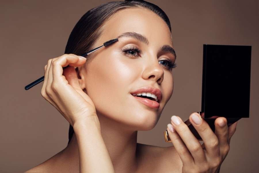 Une jeune femme souriante applique un maquillage de jour lumineux en se regardant dans un miroir de poche.
